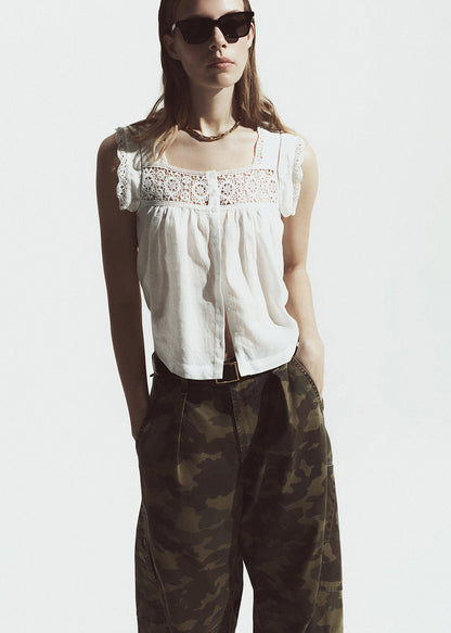 Woman wearing a white sleeveless top with lace details and camouflage pants on a plain background