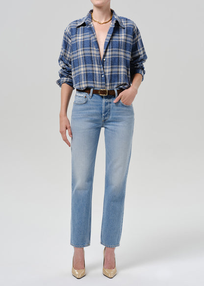 A model wearing a blue and white plaid button-down shirt tucked into light-wash blue jeans, styled with a brown belt, gold necklace, and gold high heels against a grey background.