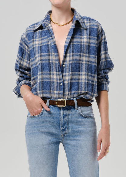 A person wearing a blue and beige plaid button-up shirt, light-wash blue jeans, a brown belt with a gold buckle, and a gold chain necklace, photographed against a grey background.