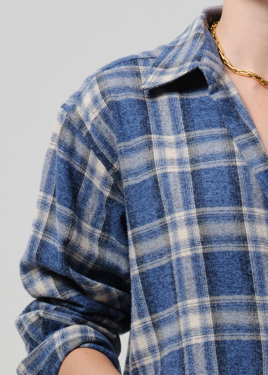 A close-up of a person wearing a blue and off-white plaid button-up shirt, with a gold chain necklace visible against a grey background.