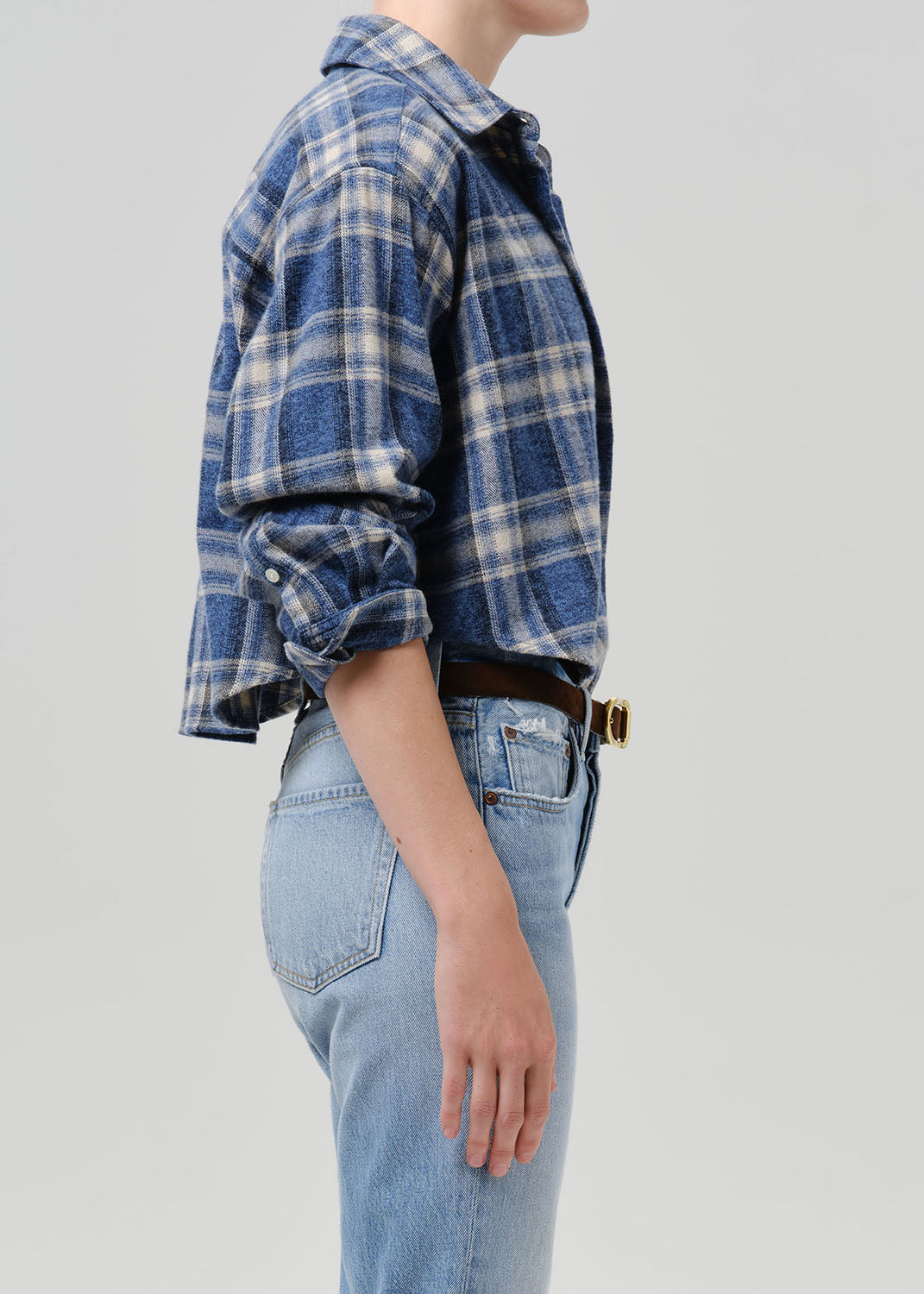 Side profile of a person wearing a blue and white plaid button-up shirt with light-wash blue jeans, a brown belt, and a gold buckle, against a grey background.