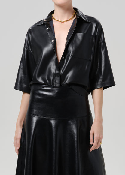 A model wearing a black faux leather short-sleeve button-up shirt and matching black faux leather skirt against a grey background.