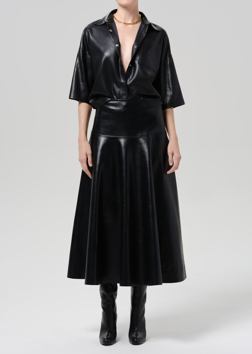A model wearing a black faux leather two-piece set consisting of a short-sleeve button-down shirt and a matching flared midi skirt, paired with black boots against a grey background.