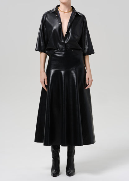 A model wearing a black faux leather two-piece set consisting of a short-sleeve button-down shirt and a matching flared midi skirt, paired with black boots against a grey background.