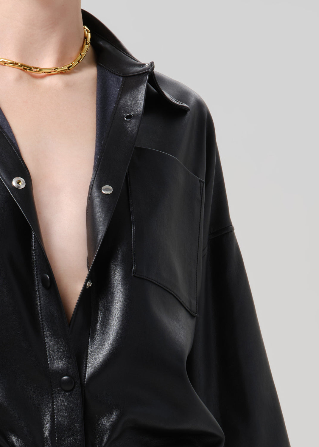 Close-up of a person wearing a black leather-look button-up shirt with a collar, chest pocket, and silver-tone buttons, paired with a chunky gold chain necklace against a grey background.