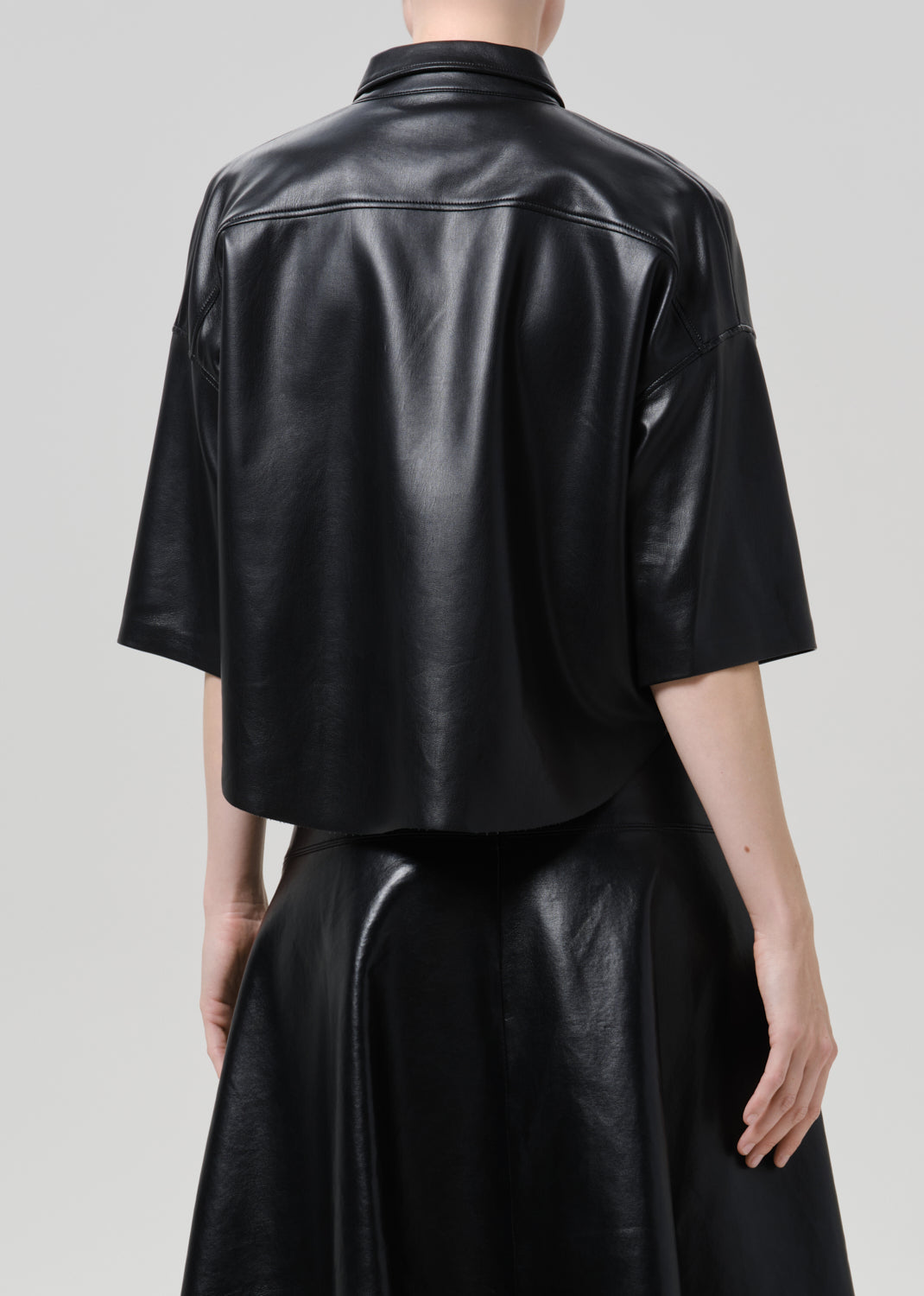 Rear view of a model wearing a black leather cropped shirt jacket with elbow-length sleeves and a matching black leather skirt against a grey background.