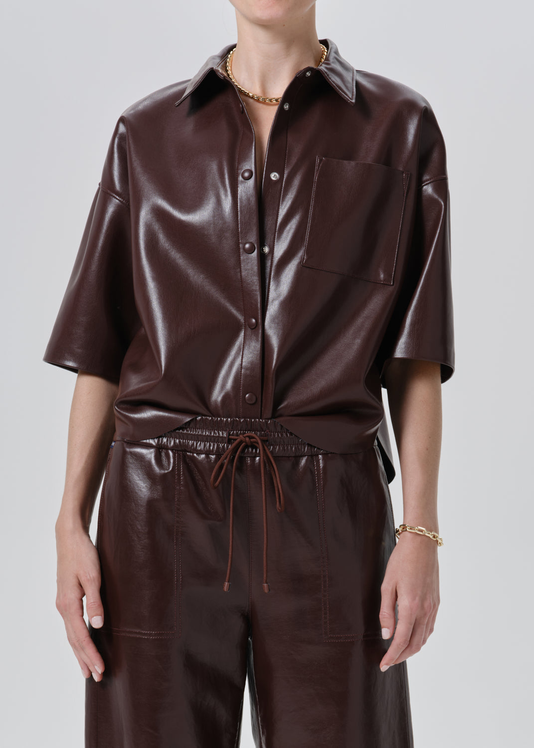 A model wearing a dark brown faux leather short-sleeve button-up shirt and matching dark brown faux leather trousers, shown from the neck down against a grey background.