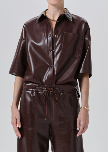 A model wearing a dark brown faux leather short-sleeve button-up shirt and matching dark brown faux leather trousers, shown from the neck down against a grey background.