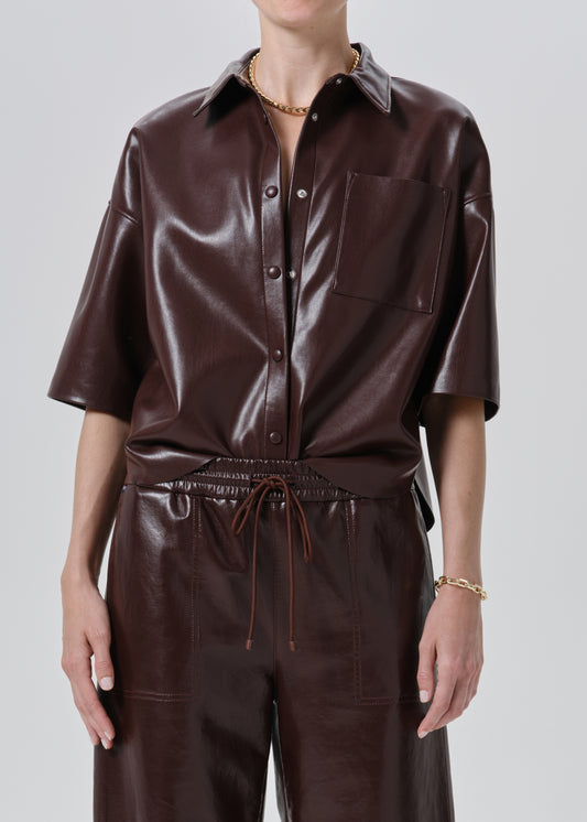 A model wearing a dark brown faux leather short-sleeve button-up shirt and matching dark brown faux leather trousers, shown from the neck down against a grey background.