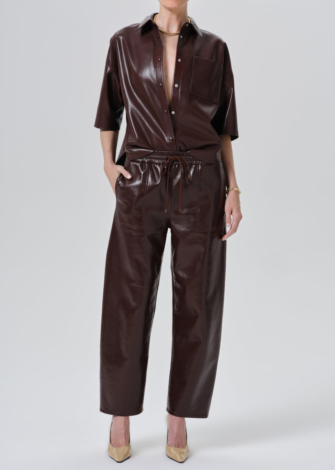 A model wearing a dark brown faux leather jumpsuit with a button-up shirt collar, short sleeves, and a drawstring waist, paired with beige pointed-toe heels against a grey background.