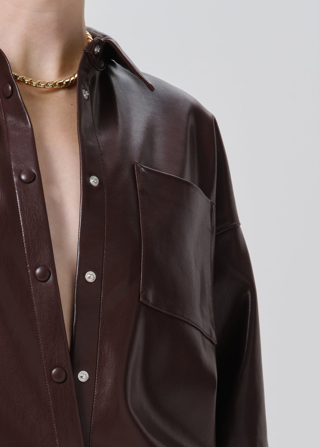 Close-up of a person wearing an oversized dark brown faux-leather button-up shirt with a chest pocket and a gold chain necklace, against a grey background.