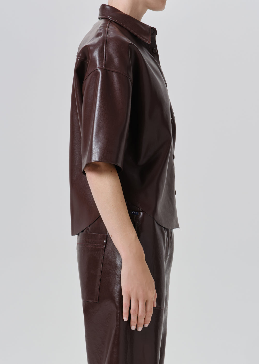 A side-profile view of a model wearing a dark brown faux-leather short-sleeve button-up shirt and matching dark brown trousers against a grey background.
