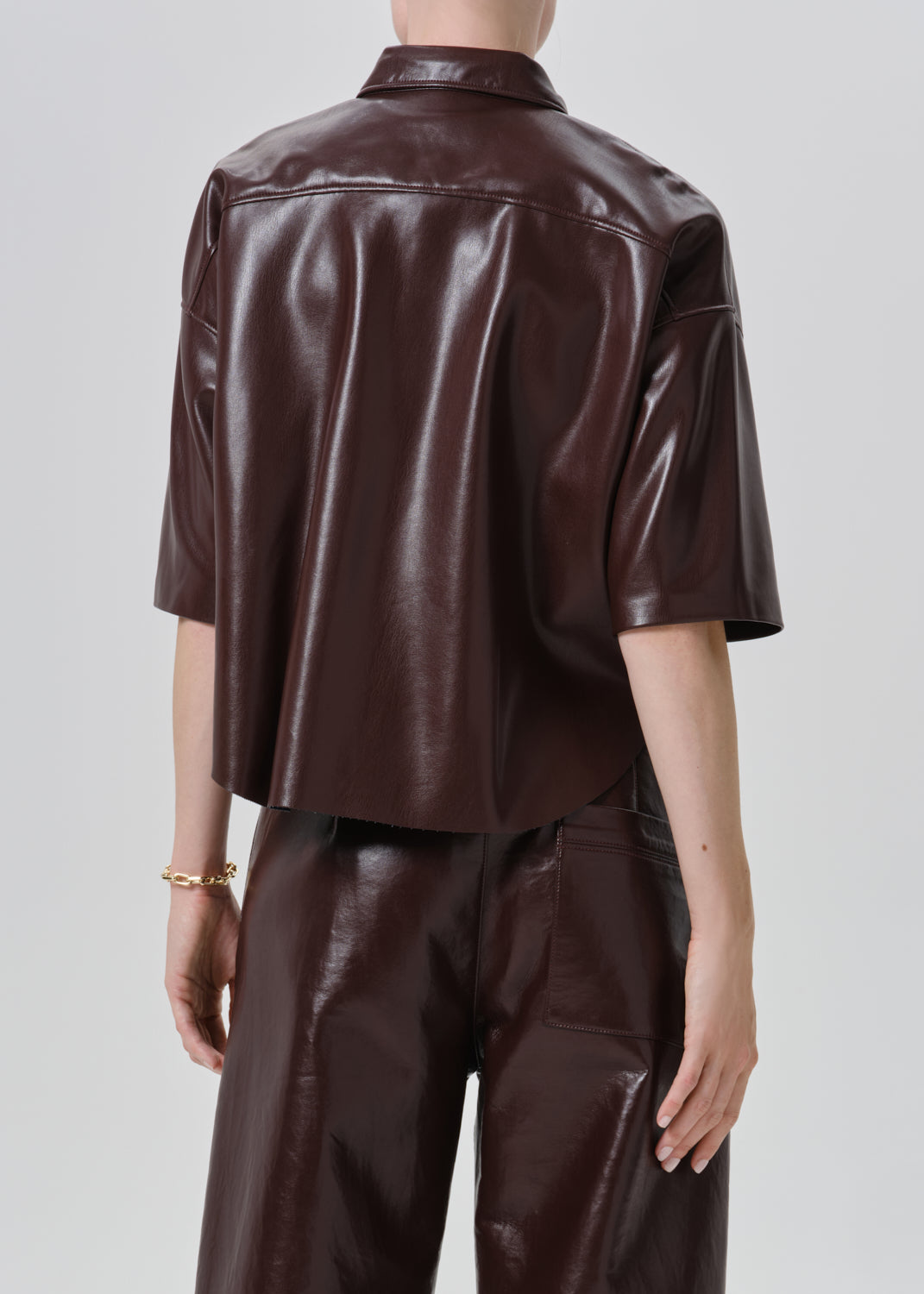 A model shown from the back wearing a dark brown faux-leather short-sleeve button-up shirt and matching dark brown trousers against a grey background.