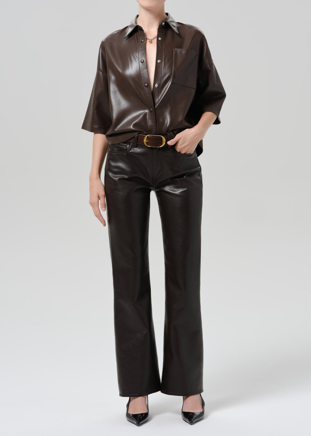 A model wearing a dark brown glossy faux-leather outfit consisting of a short-sleeve button-up shirt and matching wide-leg pants with a gold-tone belt, paired with black pointed-toe heels against a grey background.