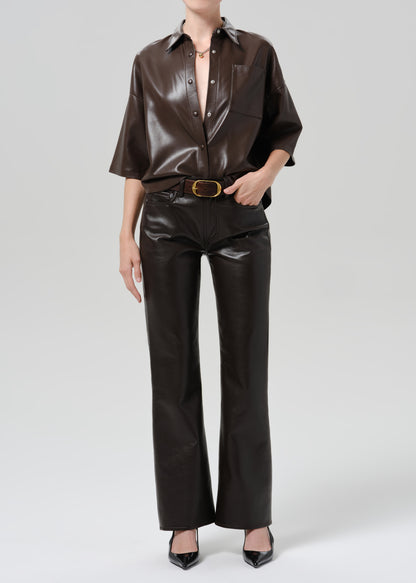 A model wearing a dark brown glossy faux-leather outfit consisting of a short-sleeve button-up shirt and matching wide-leg pants with a gold-tone belt, paired with black pointed-toe heels against a grey background.