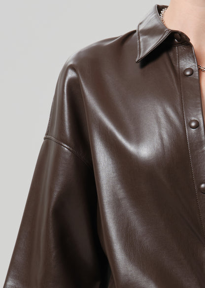 Close-up of a person wearing an oversized dark brown faux leather button-up shirt with a pointed collar and visible buttons, against a grey background.