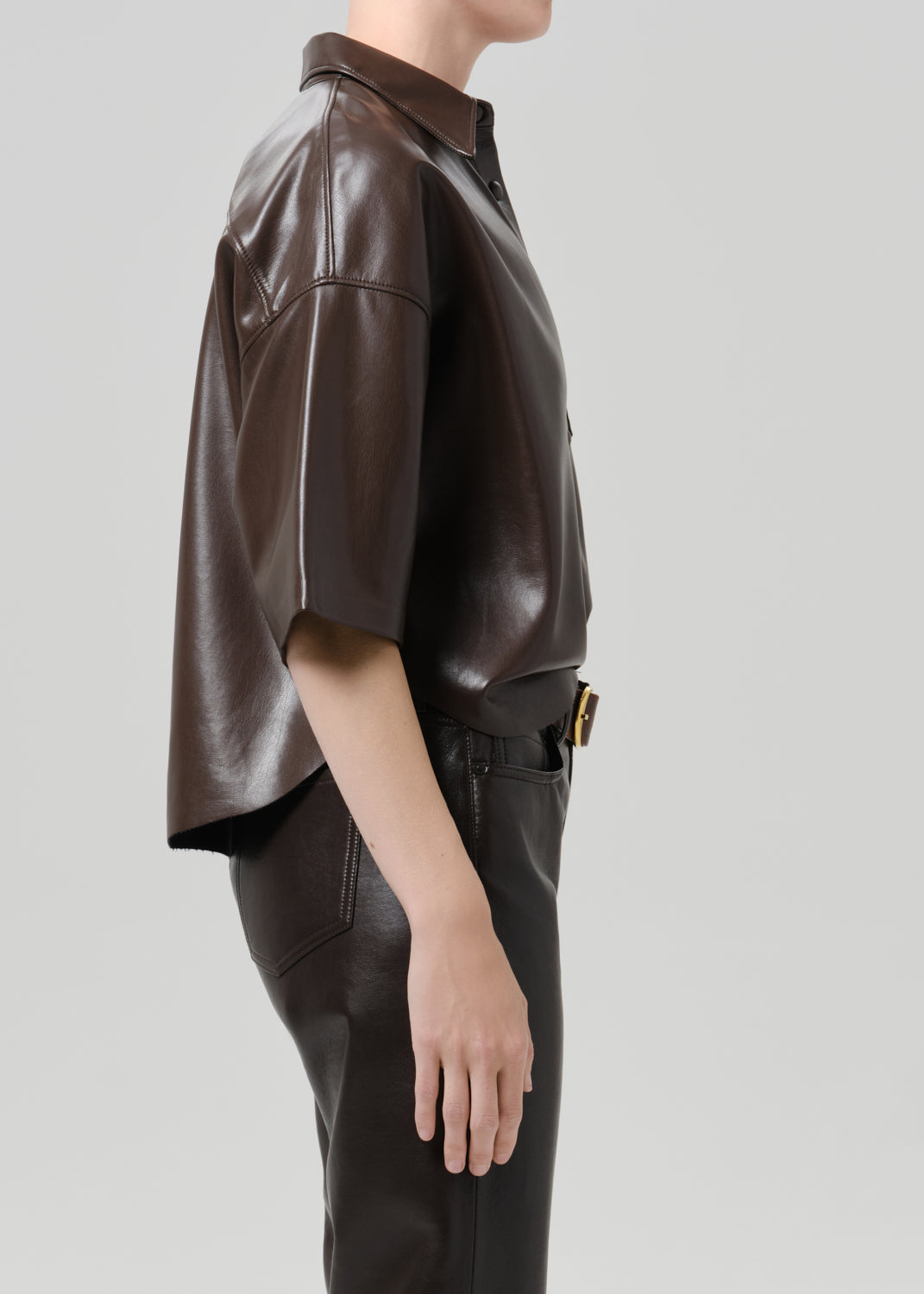 A side-profile view of a model wearing a dark brown leather short-sleeve shirt and matching dark brown leather pants against a grey background.