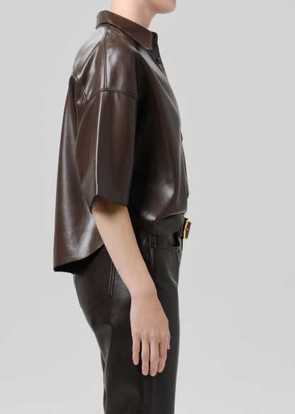 A side-profile view of a model wearing a dark brown leather short-sleeve shirt and matching dark brown leather pants against a grey background.