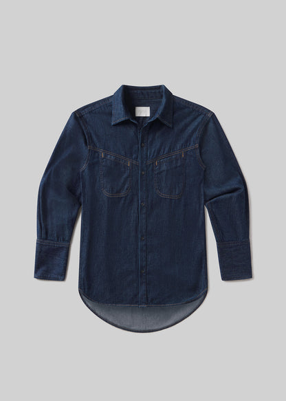 Dark indigo long-sleeve denim shirt with a pointed collar, front button closure, two chest pockets, contrast stitching, buttoned cuffs, and a curved hem, front flat lay view on a grey background