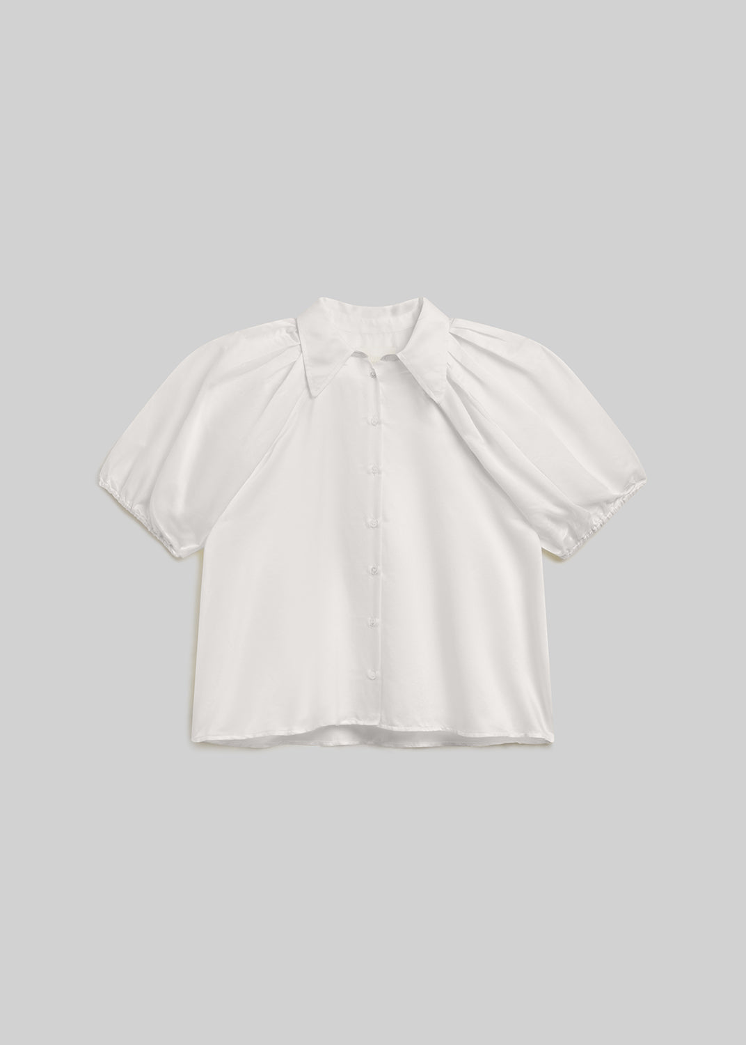White short-sleeve blouse with a pointed collar, front button closure, voluminous puff sleeves, and gathered elastic cuffs, front flat lay view on a grey background.