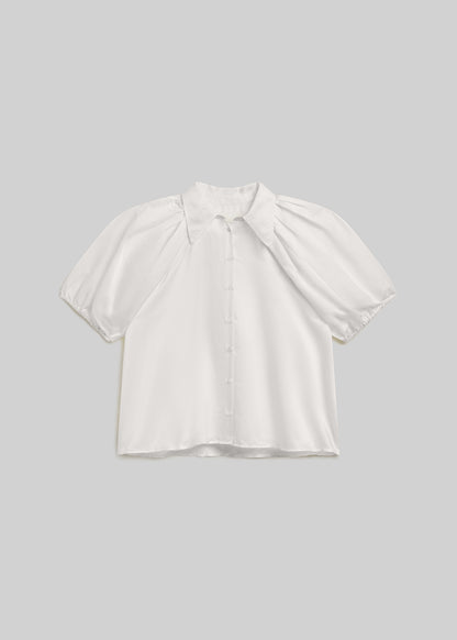 White short-sleeve blouse with a pointed collar, front button closure, voluminous puff sleeves, and gathered elastic cuffs, front flat lay view on a grey background.