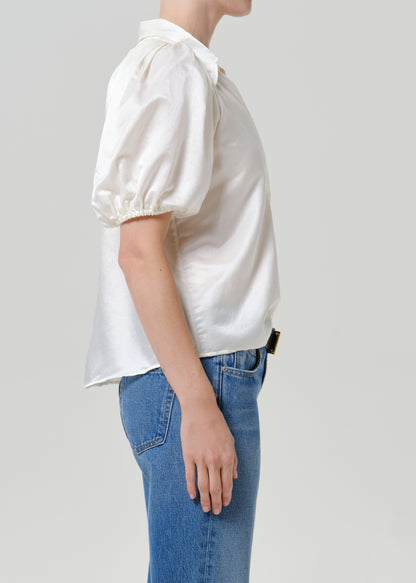 Woman wearing a white puff sleeve button-up blouse with medium blue straight leg jeans viewed from the side on a grey background.
