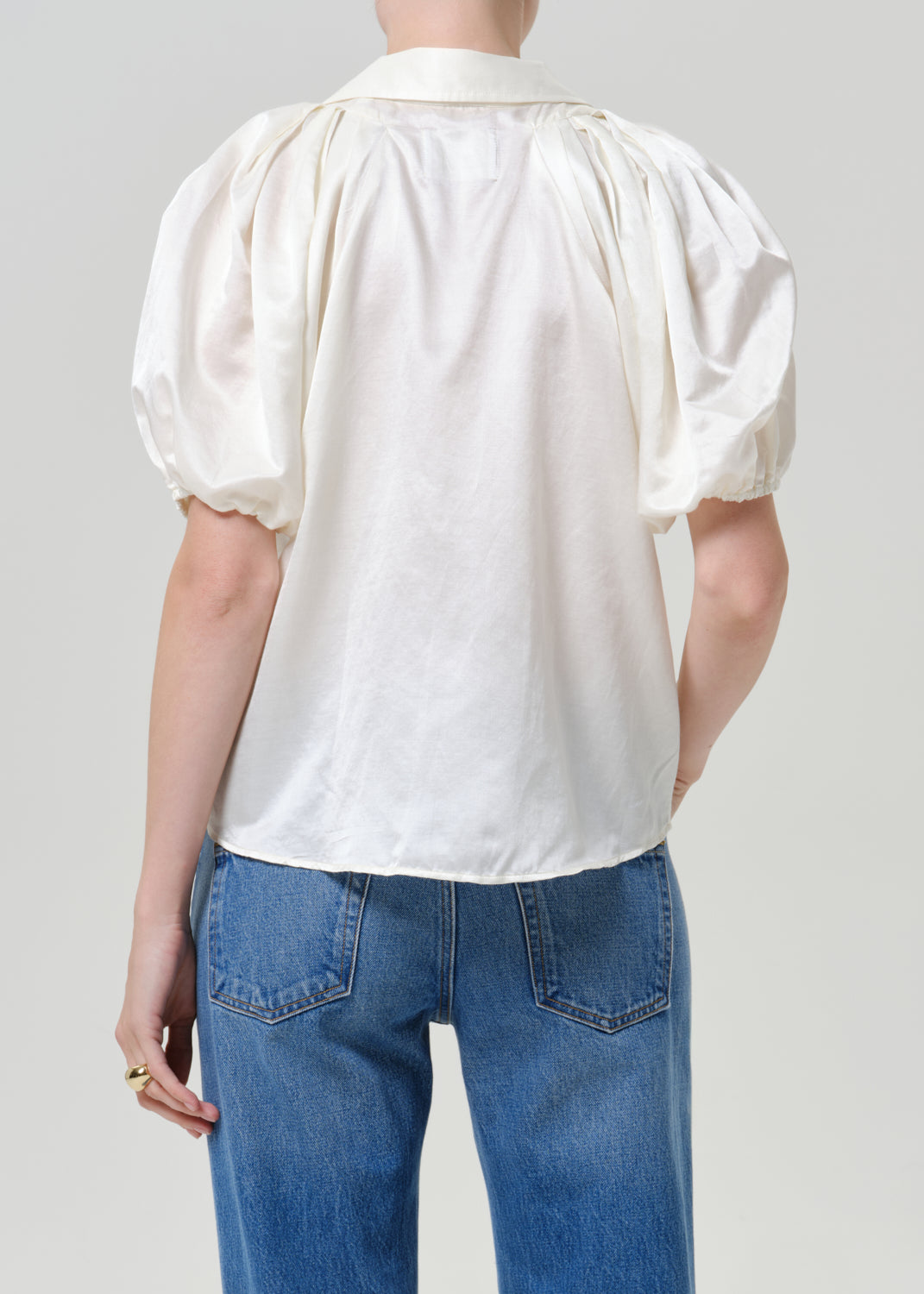 Woman wearing a white puff sleeve button-up blouse with medium blue straight leg jeans viewed from the back on a grey background.