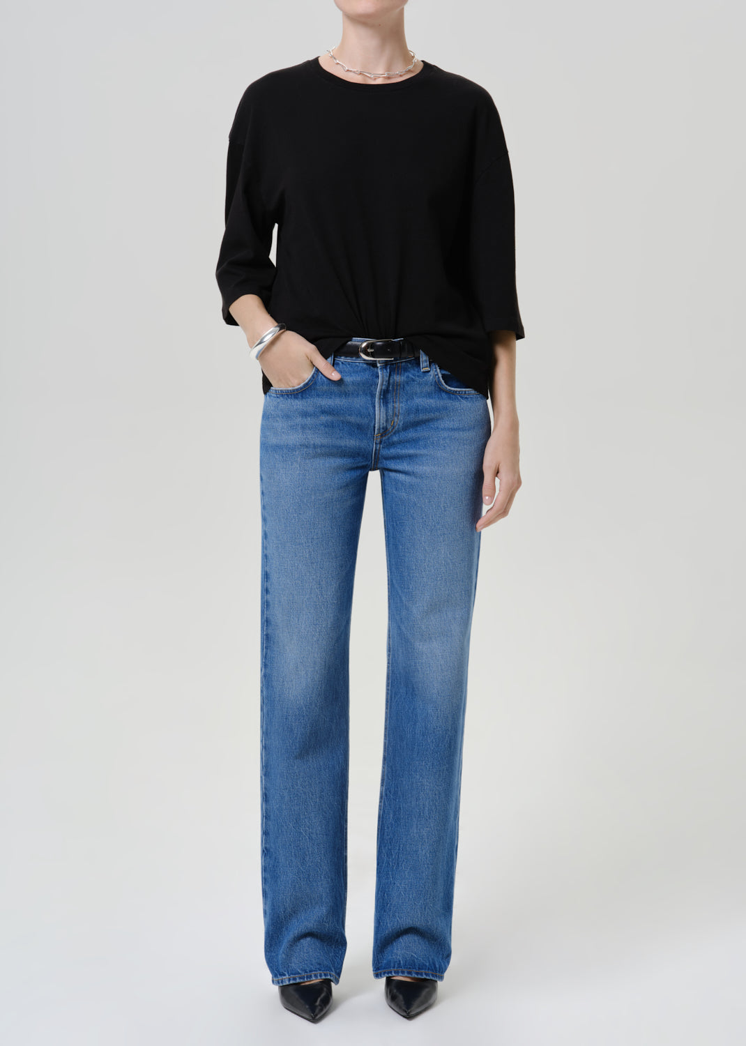 A model wearing a black long-sleeve top and blue straight-leg jeans with a black belt, standing against a light gray studio background.