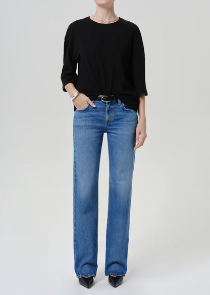 A model wearing a black long-sleeve top and blue straight-leg jeans with a black belt, standing against a light gray studio background.