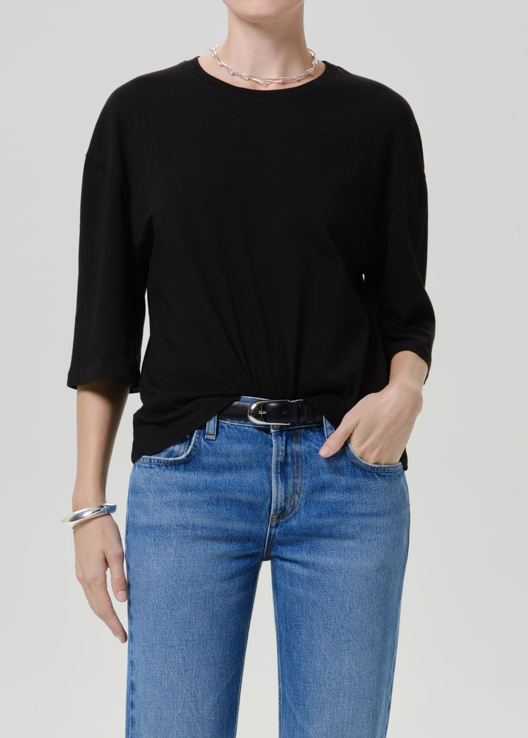 A model wearing a black three-quarter sleeve top tucked into blue jeans with a black belt, photographed against a light gray studio background.