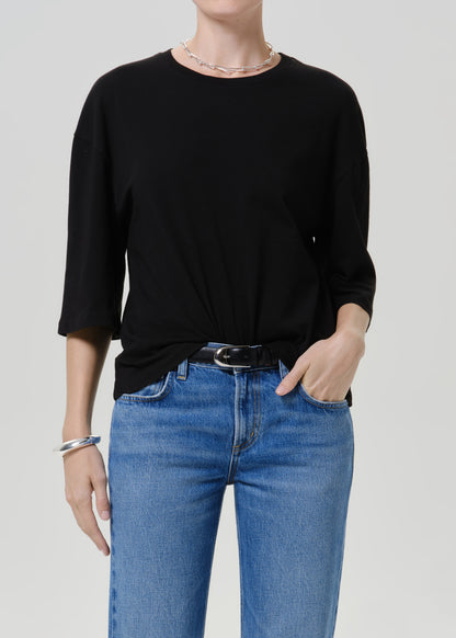A model wearing a black three-quarter sleeve top tucked into blue jeans with a black belt, photographed against a light gray studio background.