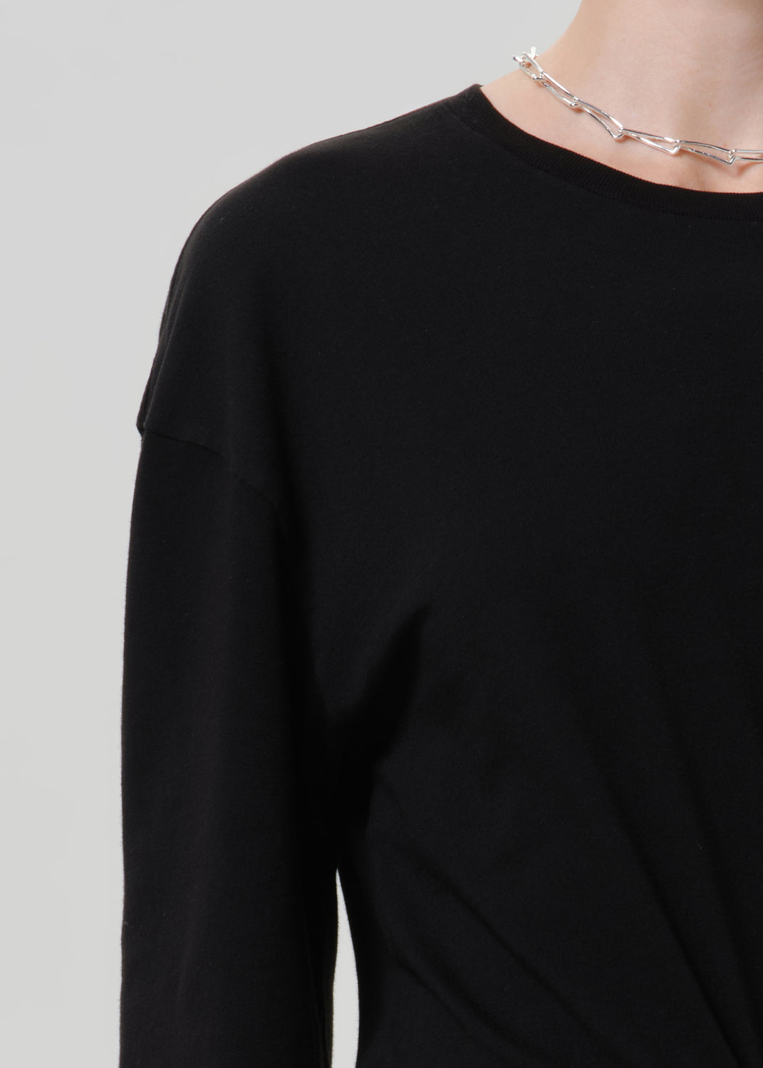 Close-up of a person wearing a plain black long-sleeve top with a silver chain necklace, against a light gray studio background.