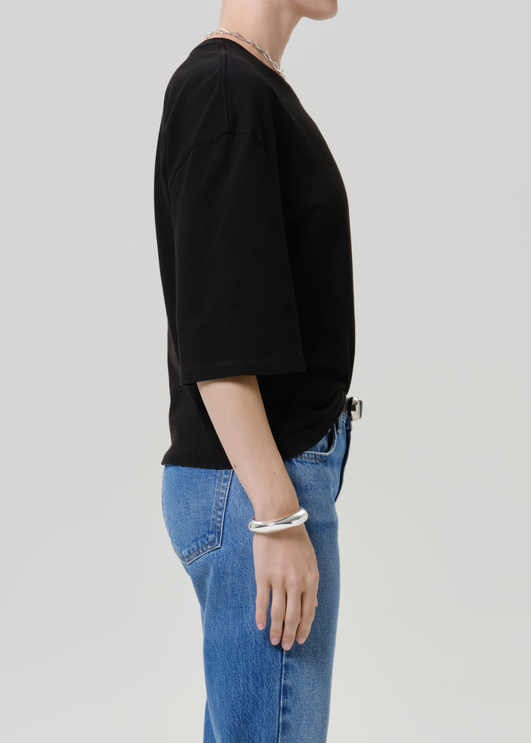 Side profile of a model wearing a black oversized short-sleeve t-shirt and blue jeans, accessorized with a silver bracelet and necklace, against a light gray studio background.