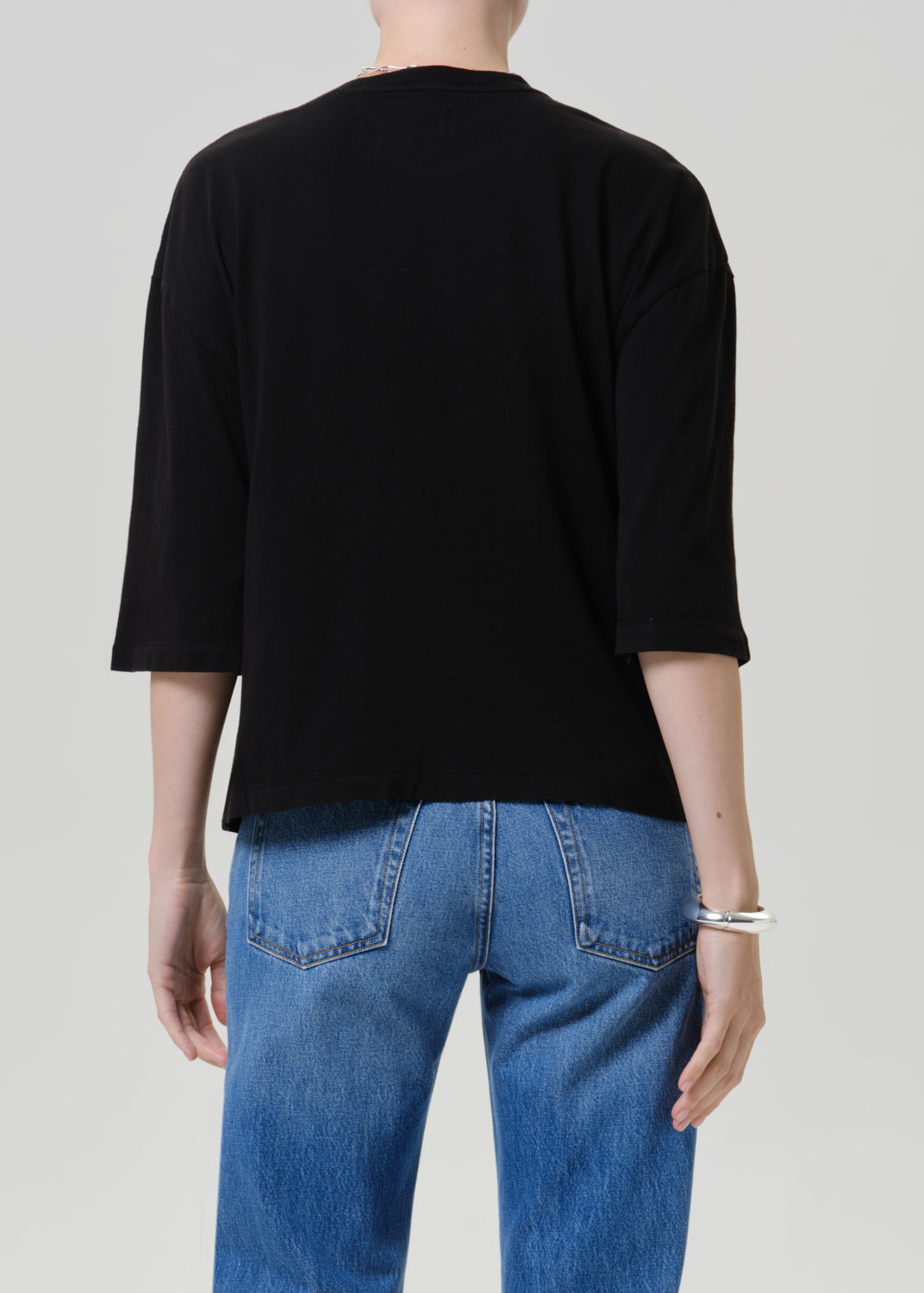 A person shown from the back wearing a plain black short-sleeve top and blue jeans against a light gray studio background.
