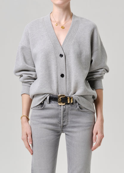 Person wearing a grey cardigan and grey jeans with a gold belt on a grey background