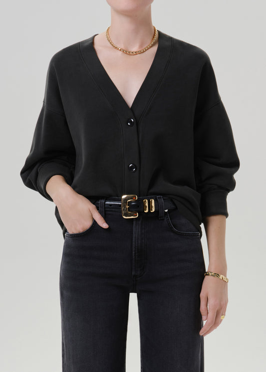 Person wearing a black long sleeve V-neck button-front cardigan with dark charcoal jeans and a black belt with gold buckle, front cropped view on a grey background.