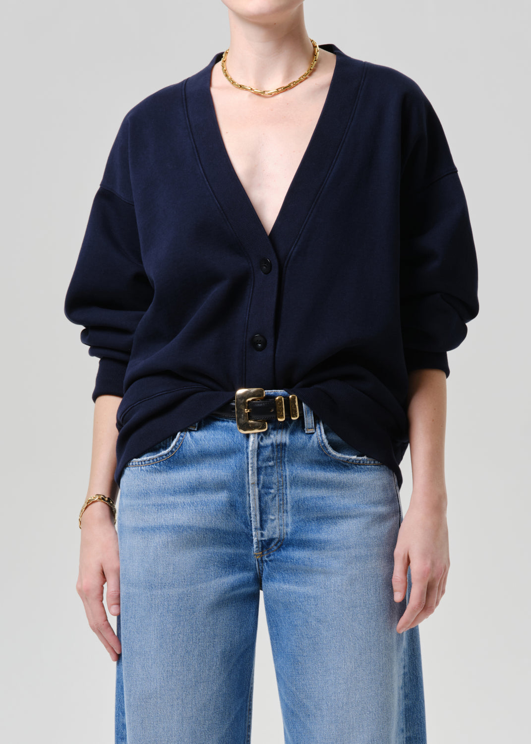 A model wearing a navy blue button-up cardigan sweater, styled with light blue jeans, a gold belt, and gold jewelry against a grey background.