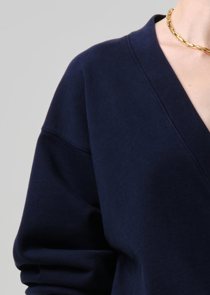 Close-up of a person wearing a dark navy blue V-neck long-sleeve top, with a gold chain necklace visible against a grey background.