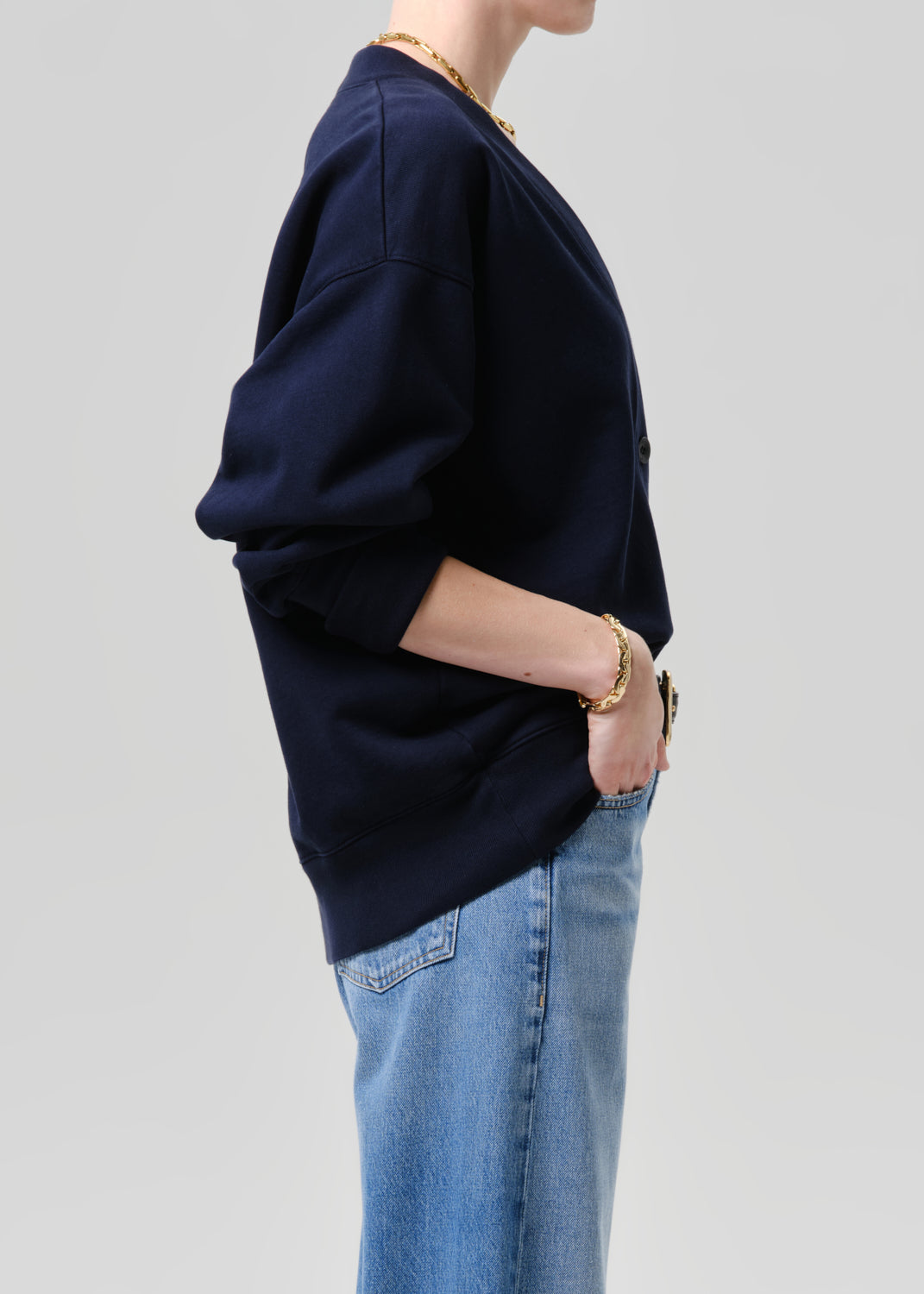 A side-profile view of a person wearing an oversized navy blue sweatshirt and light blue jeans, accessorized with gold jewelry, against a grey background.