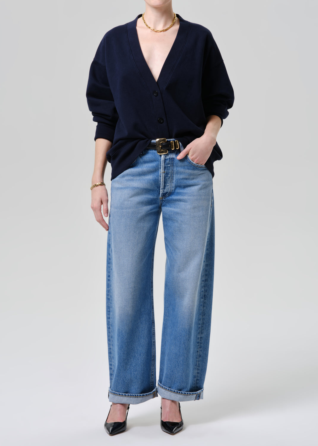 A model wearing a navy blue cardigan, light-wash wide-leg blue jeans with a black belt, and black pointed-toe heels, photographed against a grey background.