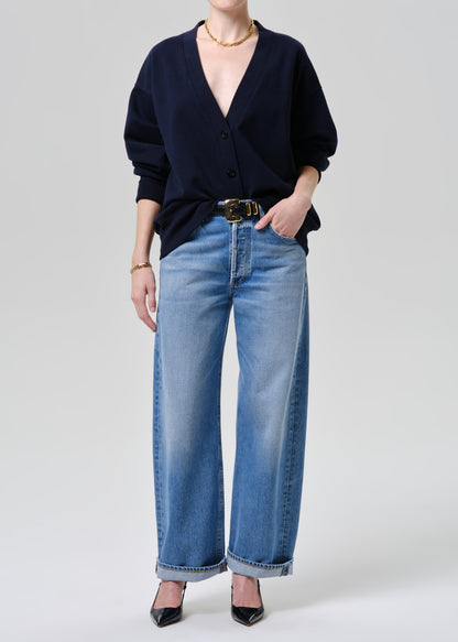 A model wearing a navy blue cardigan, light-wash wide-leg blue jeans with a black belt, and black pointed-toe heels, photographed against a grey background.
