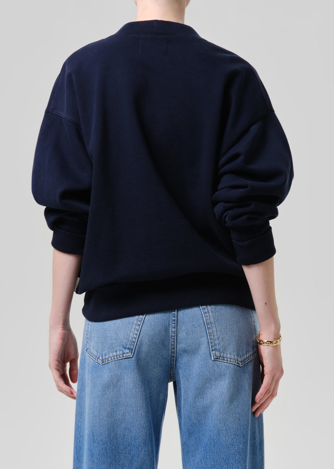 A person shown from the back wearing a dark navy crewneck sweatshirt and light blue jeans against a grey background.