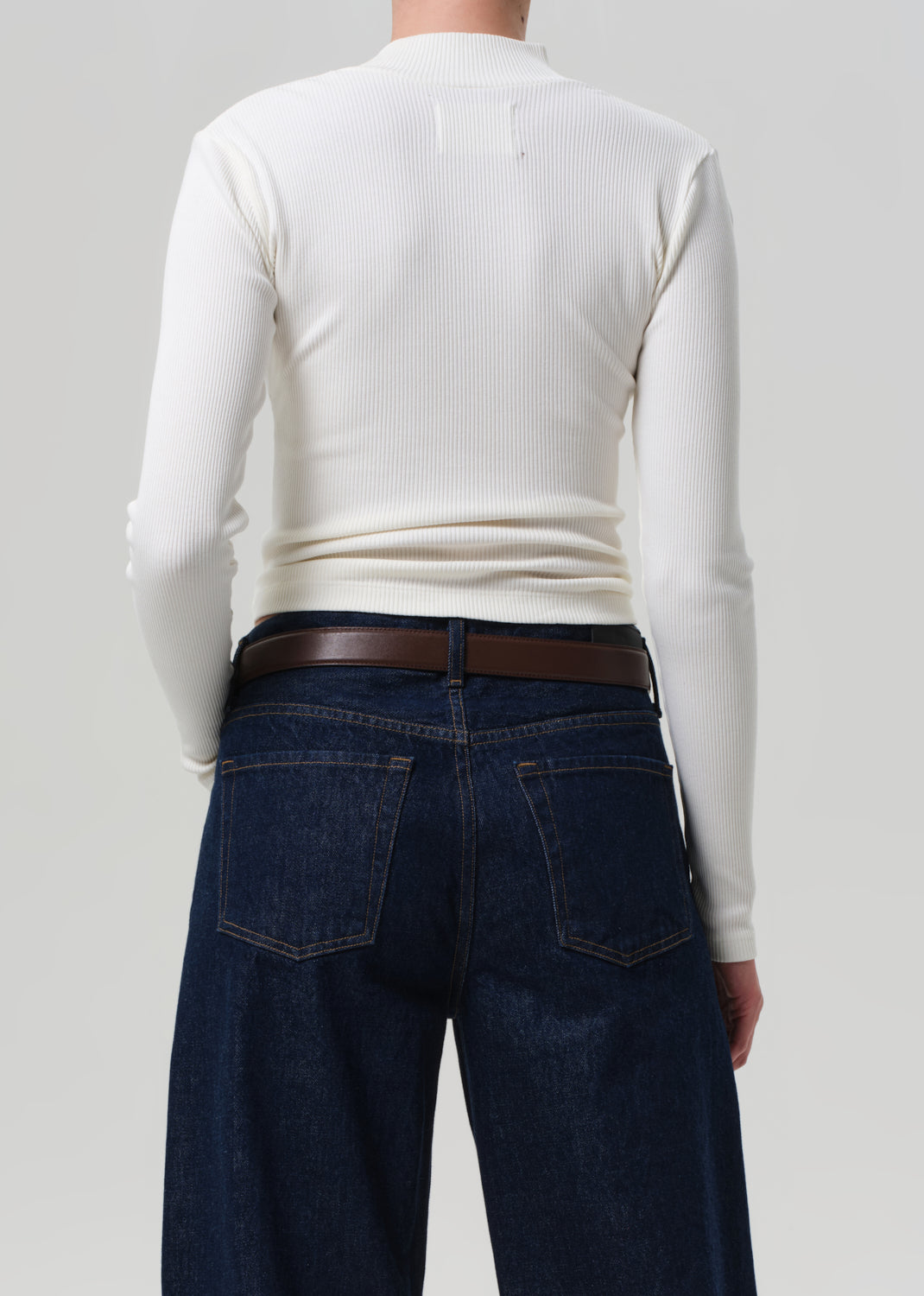 Person wearing a white long-sleeve shirt and dark blue jeans with a brown belt on a plain background