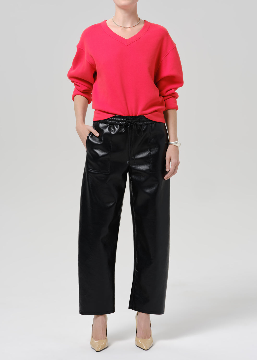 Person wearing a bright pink V neck sweater with black leather pants and beige pointed toe heels on a grey background