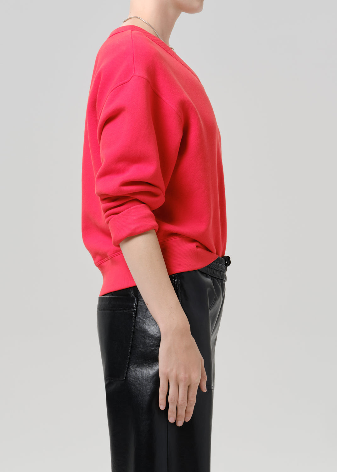 Person wearing a bright pink V neck sweater with black leather pants viewed from the side on a grey background