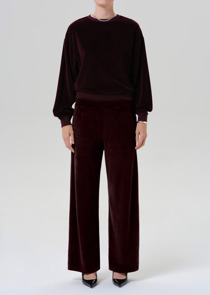 A model wearing a matching deep burgundy velvet lounge set, consisting of a long-sleeve crewneck sweatshirt and wide-leg trousers, paired with black pointed-toe heels against a grey background.