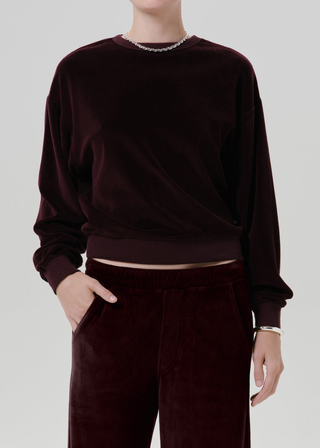 A person wearing a dark burgundy velvet long-sleeve cropped sweatshirt and matching velvet trousers, shown from the neck to the upper thighs against a grey background.