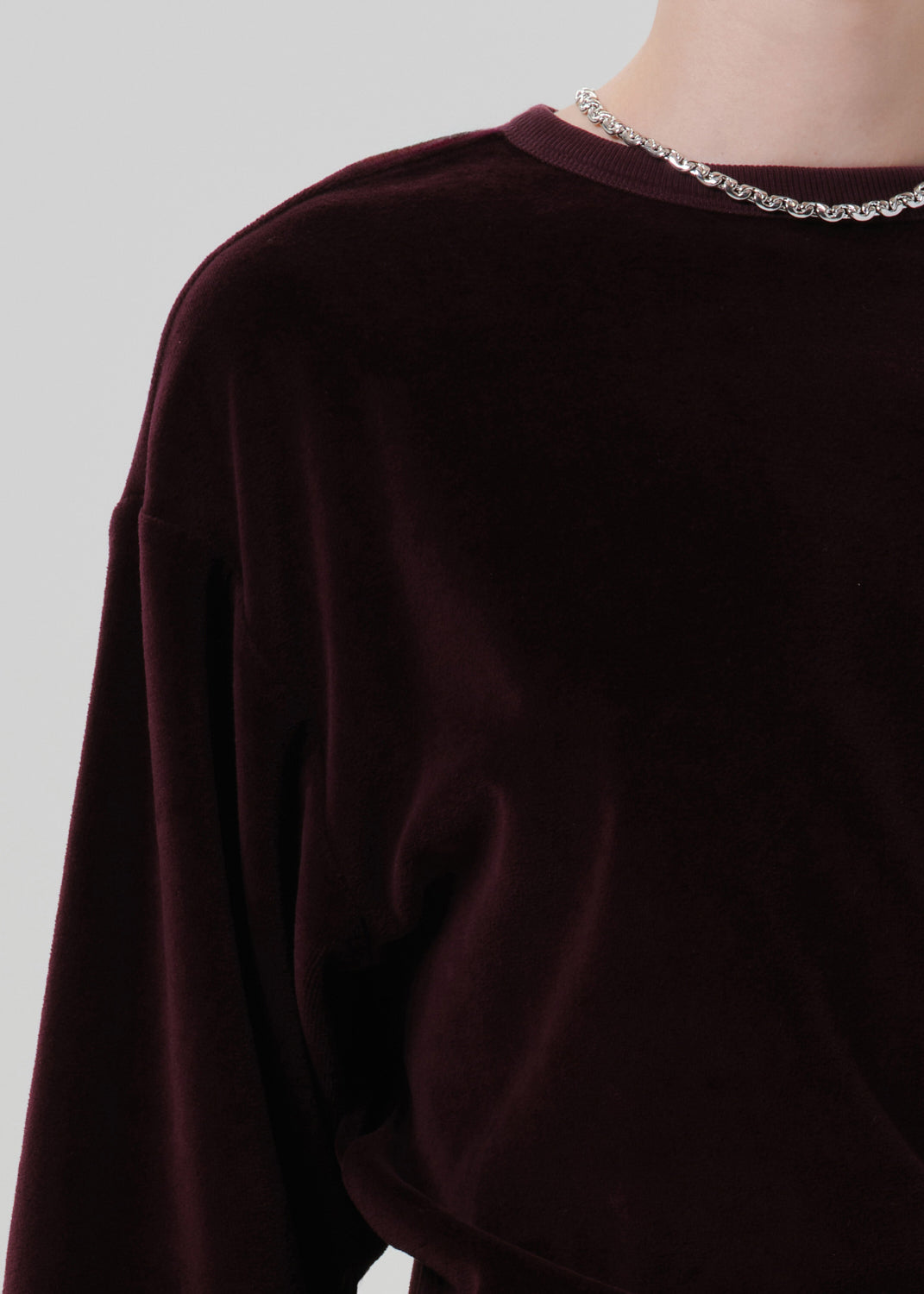Close-up of a person wearing a deep burgundy velvet long-sleeve top with a silver chain necklace, against a grey  background.