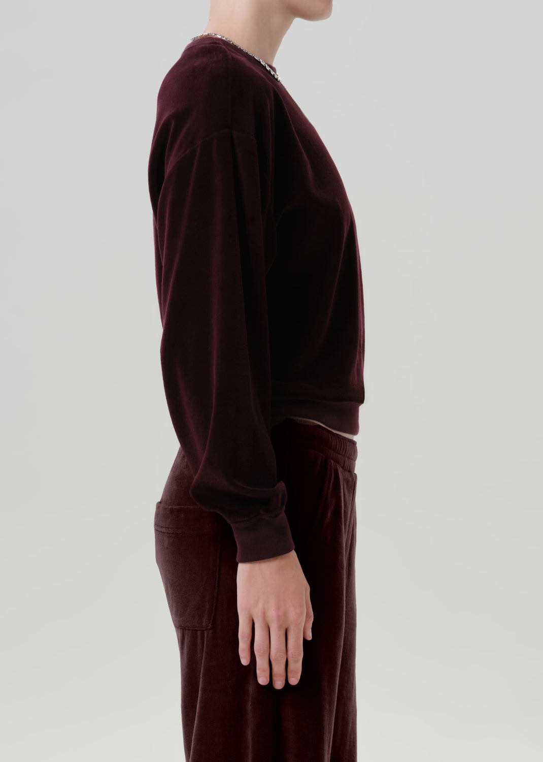 A side-profile view of a model wearing a matching deep burgundy velvet lounge set, featuring a long-sleeve top and wide-leg trousers, against a grey background.