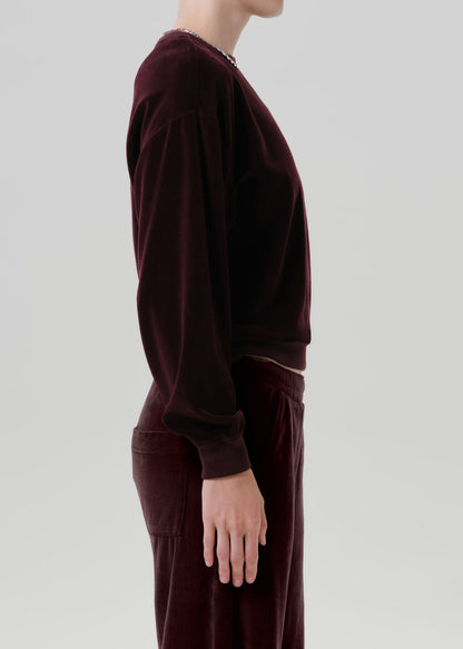 A side-profile view of a model wearing a matching deep burgundy velvet lounge set, featuring a long-sleeve top and wide-leg trousers, against a grey background.